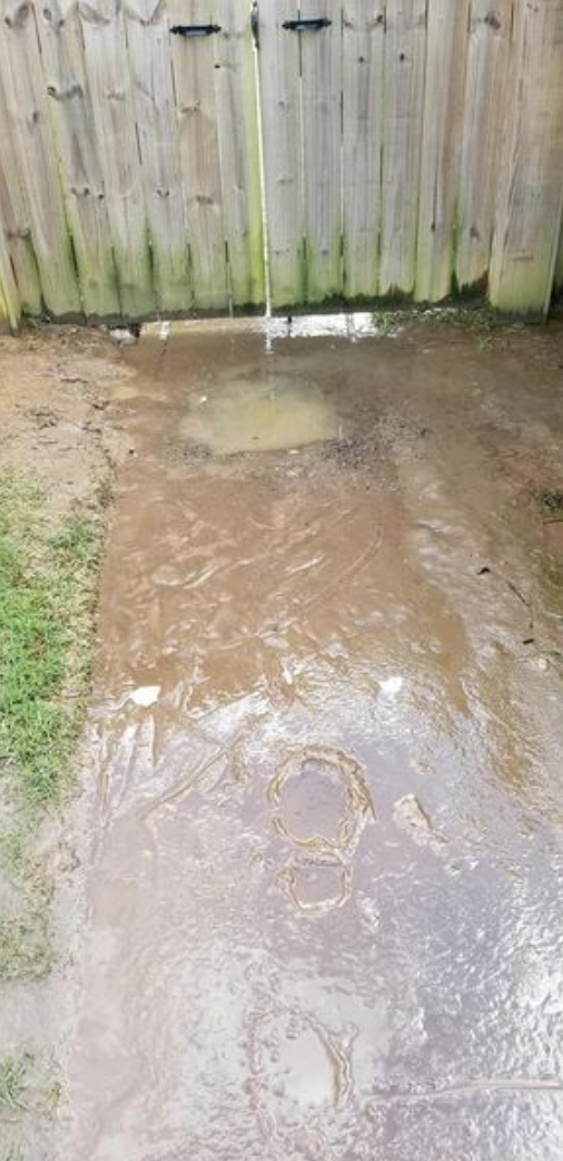 Waterlogged yard near a wooden fence with a puddle and wet concrete. Green grass on the left side.