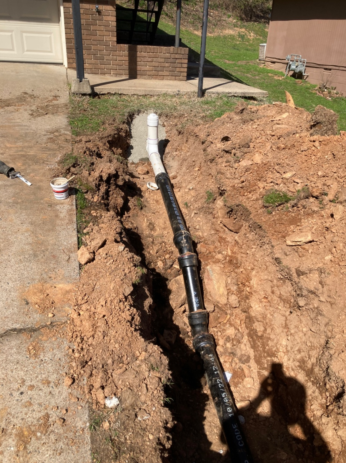 Trench in front yard with exposed pipes; a white and black pipe runs to the driveway.