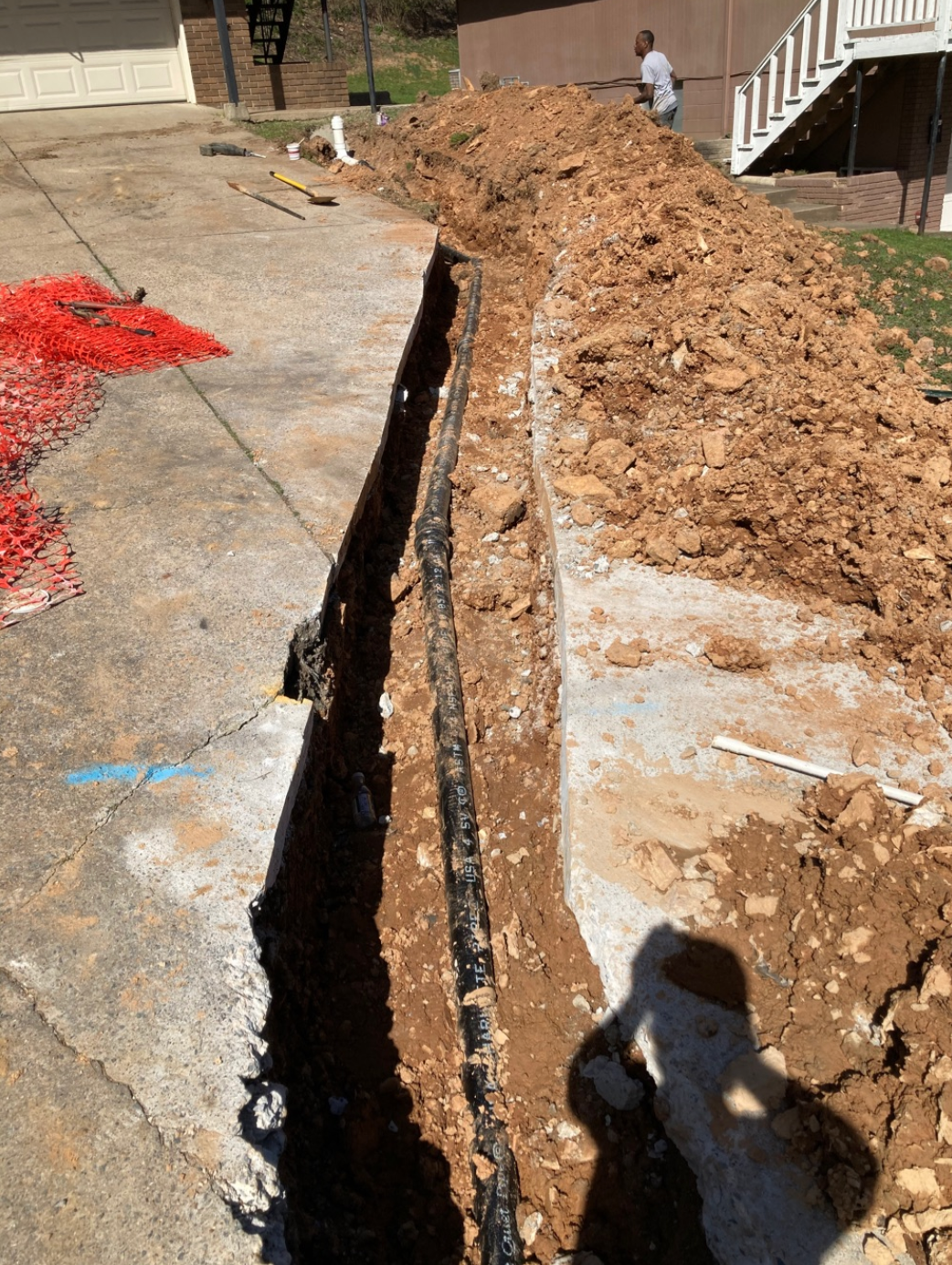 Trench dug in driveway with black pipe inside. Person in background, dirt pile on right, orange barrier left.