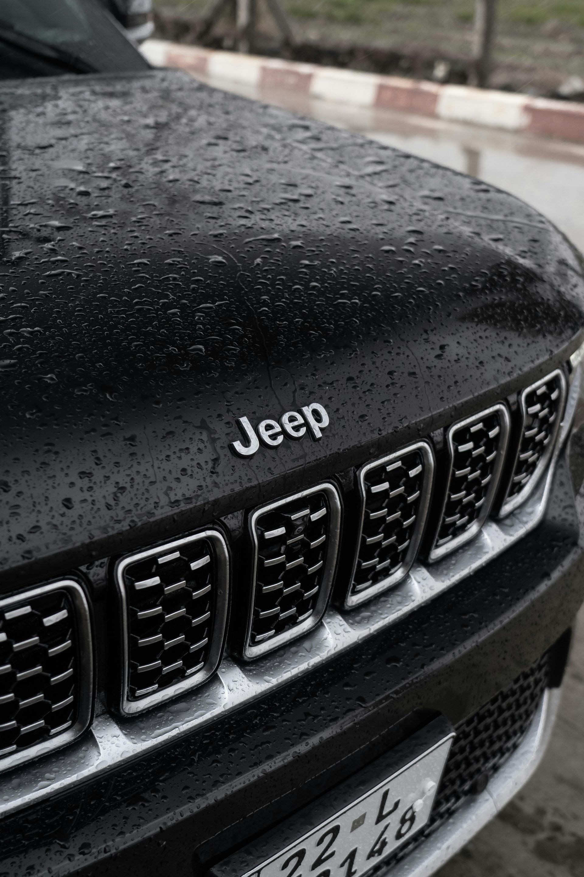 Close-up of a wet black Jeep front grille and hood with raindrops on a roadside.