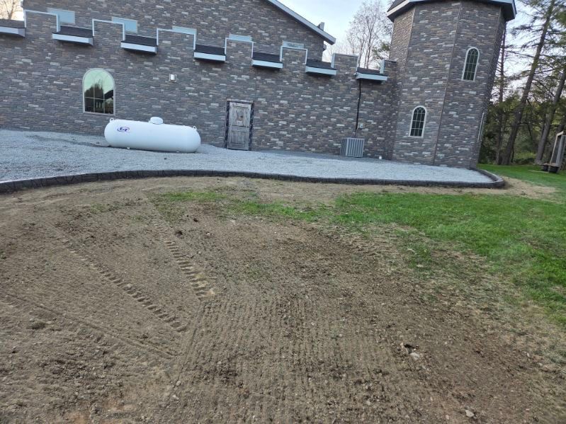 Freshly graded dirt in a yard beside a stone house and tower, with grass and a white tank nearby