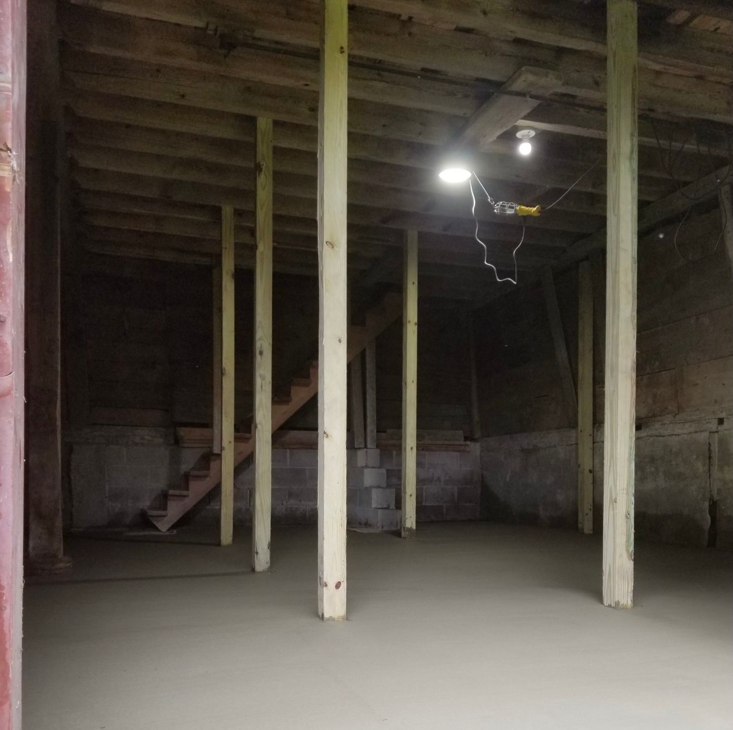Basement with concrete floor, wooden support beams, and stairs in the background. Overhead lighting.