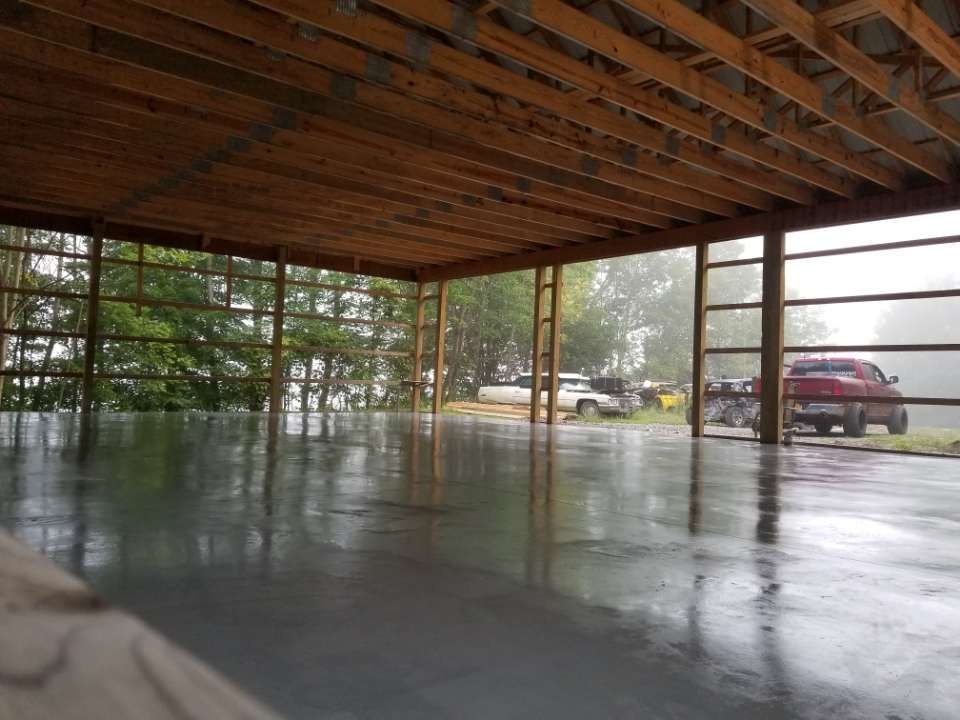 Indoor view of a concrete floor under a wooden roof. Metal beams frame the open side. Cars and trees are outside.