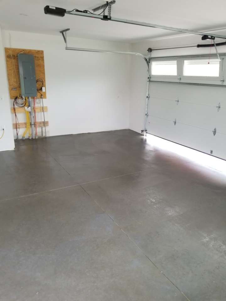 Empty garage with gray concrete floor, white walls, and open garage door. Electrical panel visible on the left wall.