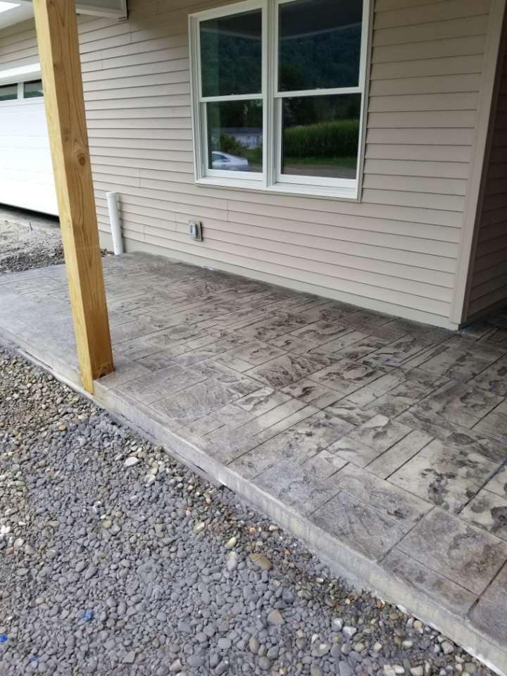 Concrete porch with wood-like texture. Beige house siding, window, and gravel path. Wooden post supports the porch roof.