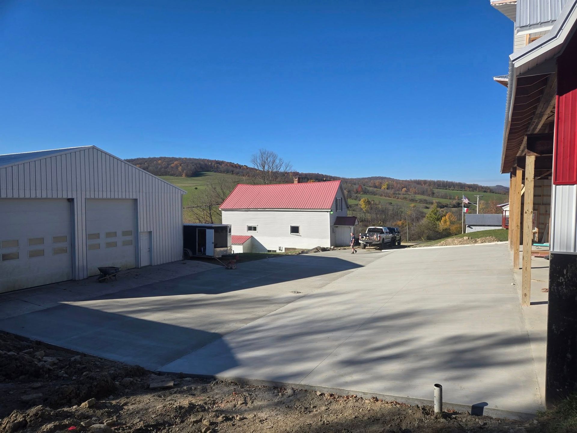 Farm scene: white garage and red-roofed building on a concrete lot, blue sky, hills in background.