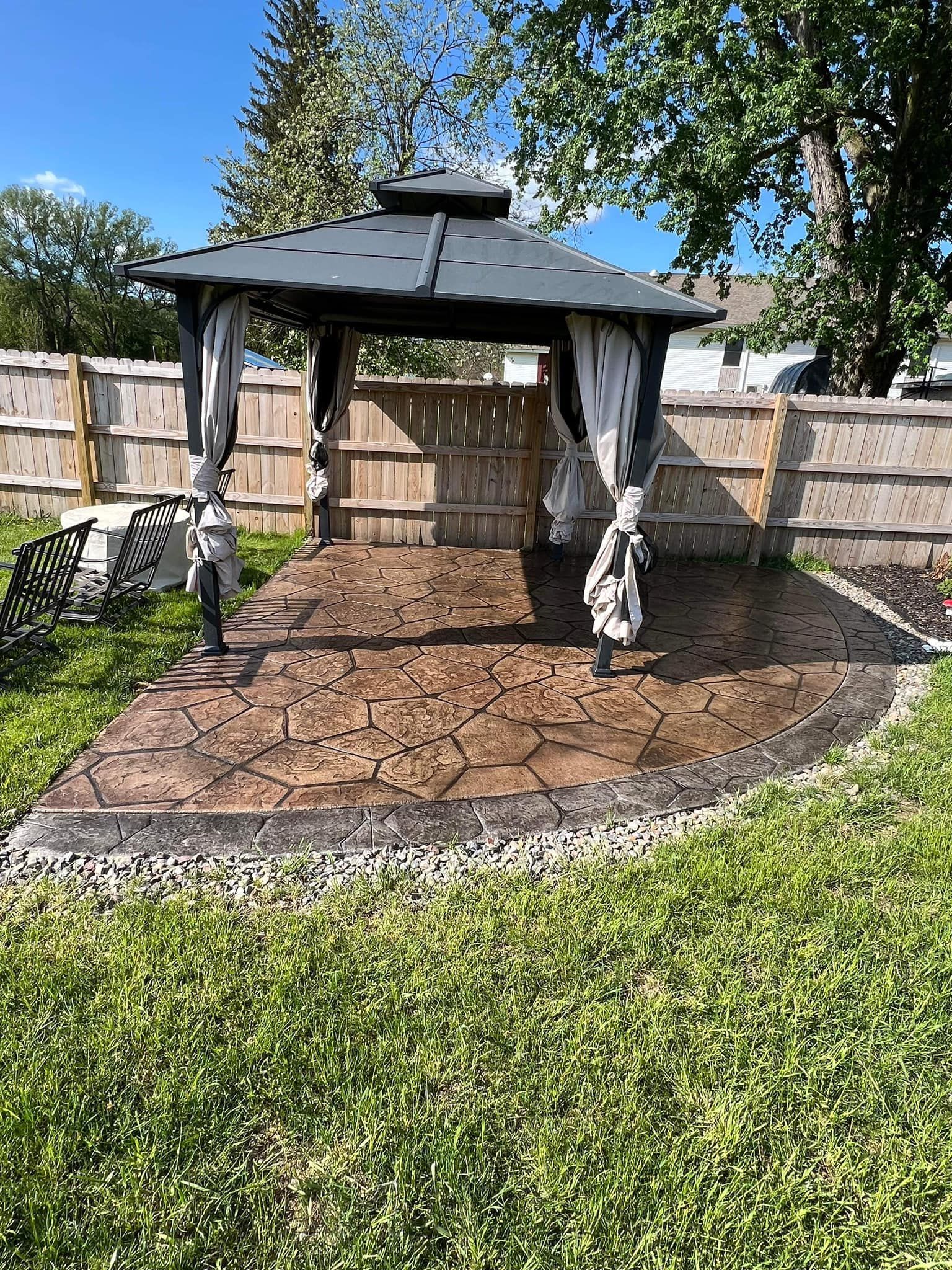Gazebo with gray roof and curtains on a stamped concrete patio, next to a wooden fence.
