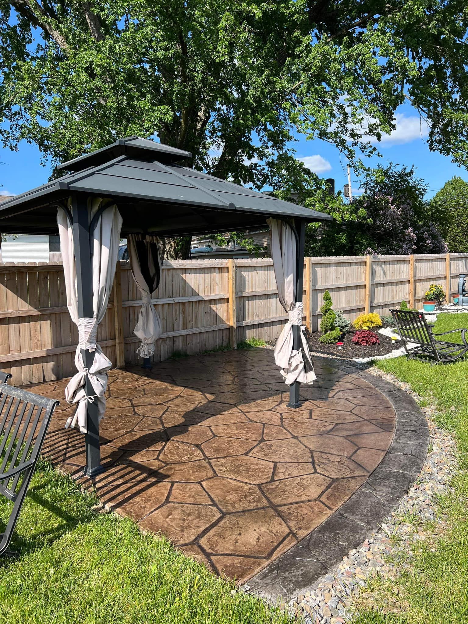 Gazebo on a stamped concrete patio in a backyard, surrounded by a wooden fence, grass, and trees.