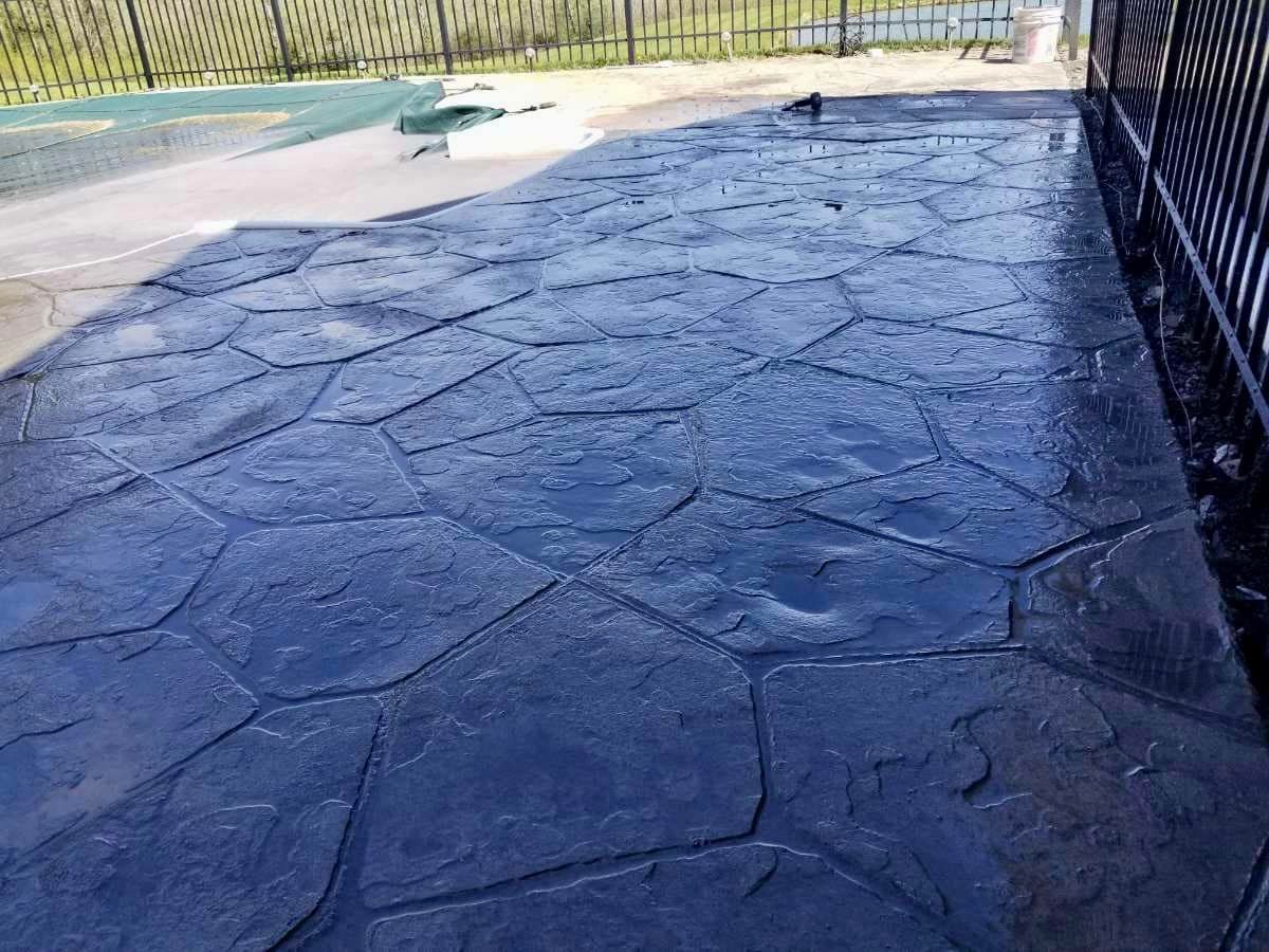 Stamped concrete patio in a dark blue, stone-like pattern, next to a black metal fence.