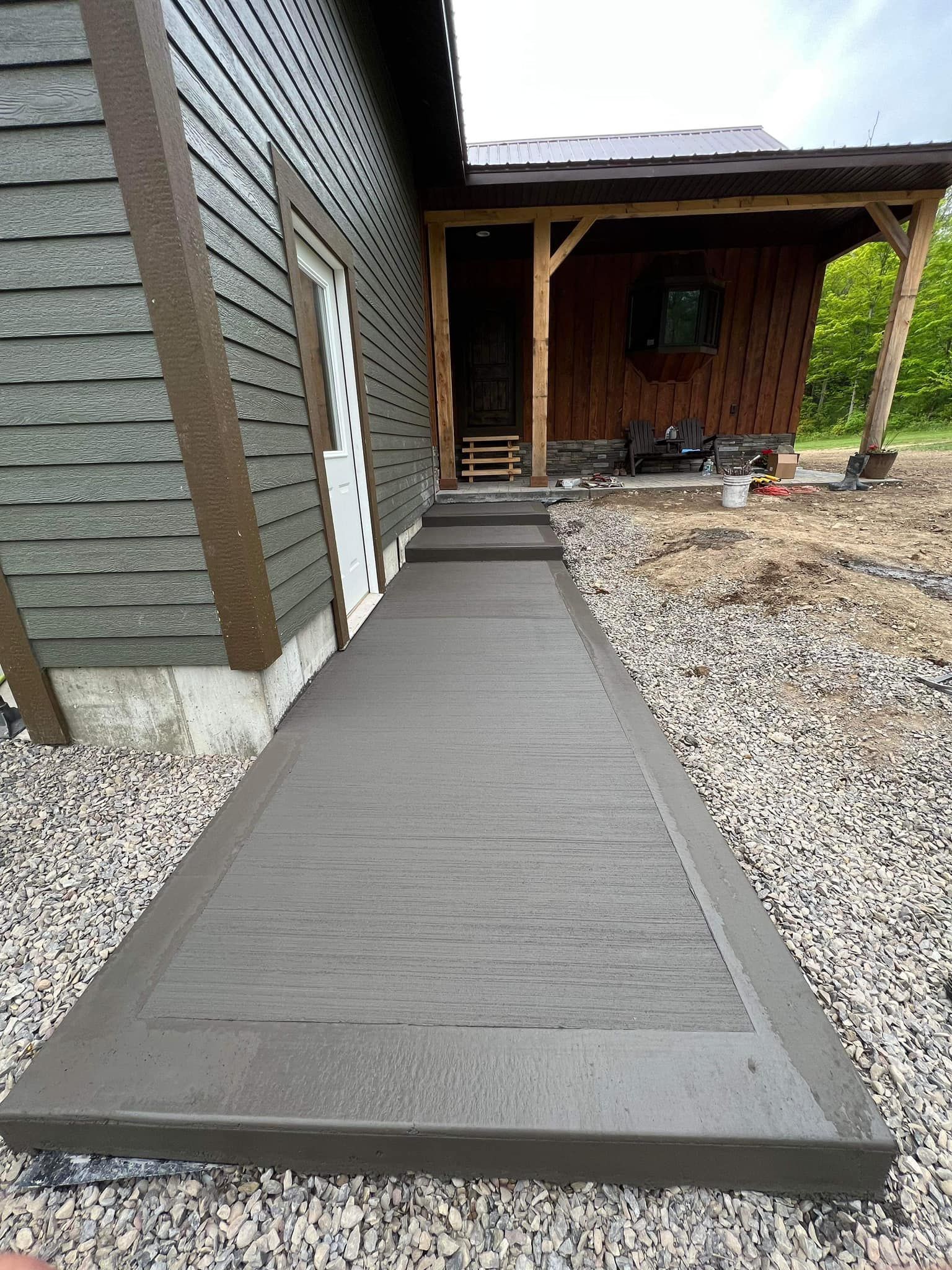 Concrete walkway with steps leading to a cabin entrance. Dark gray concrete contrasts with gravel and wooden structure.