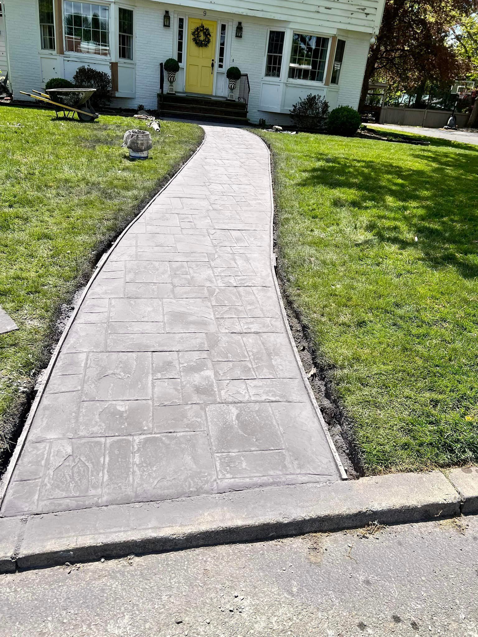 Stone walkway leading to a house with a yellow door, bordered by grass and a curb.