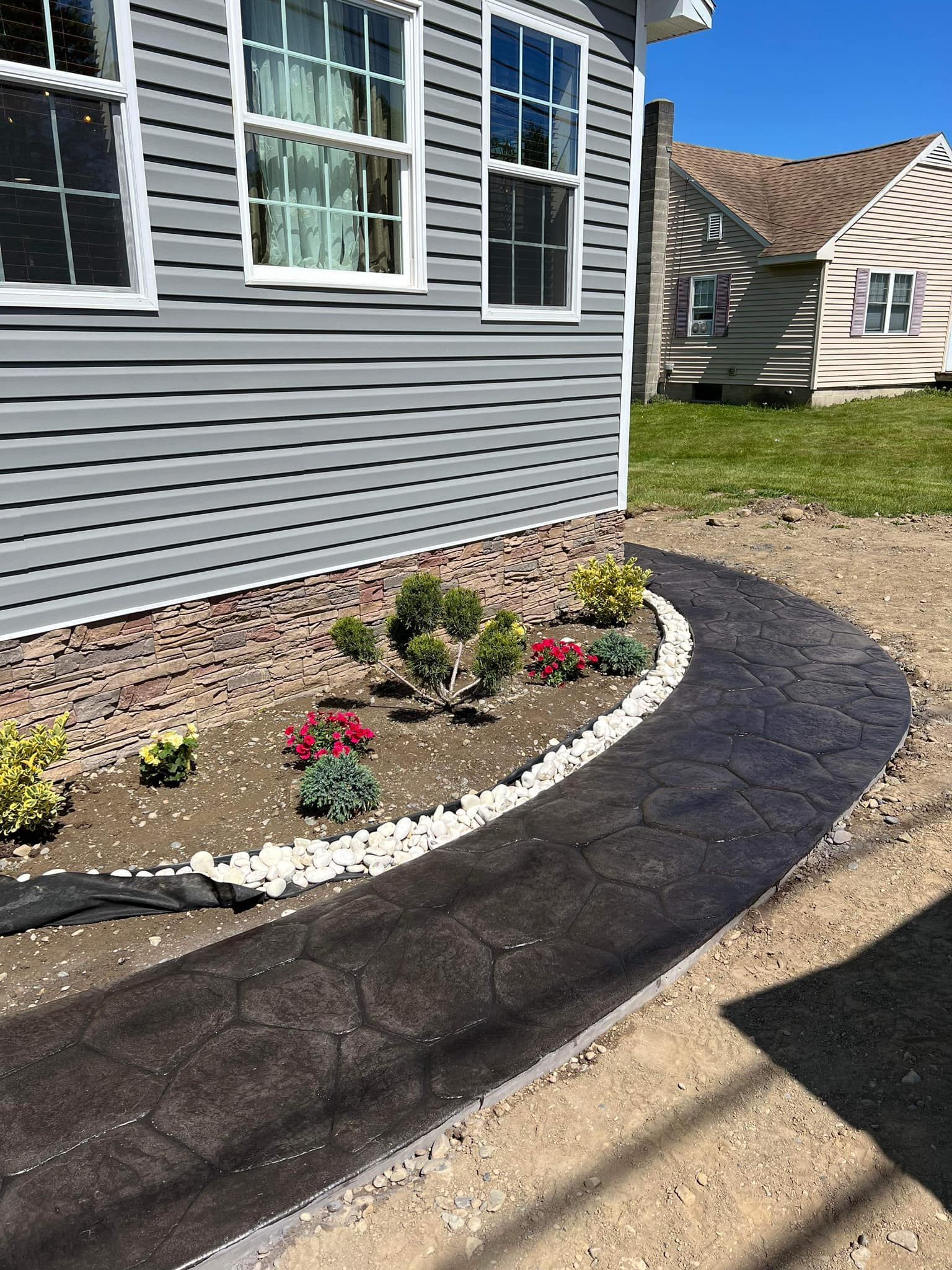Gray house with a dark patterned walkway and a small flower bed with plants and white stones.