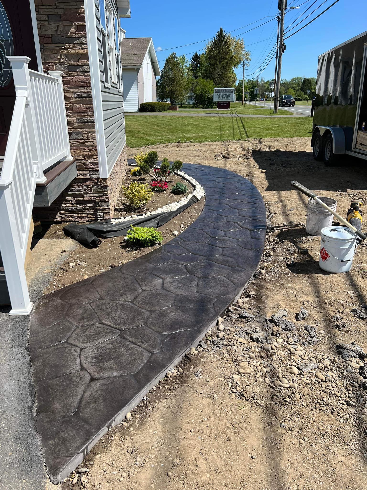 New concrete walkway leading to a house with a small flower bed alongside; sunny day.