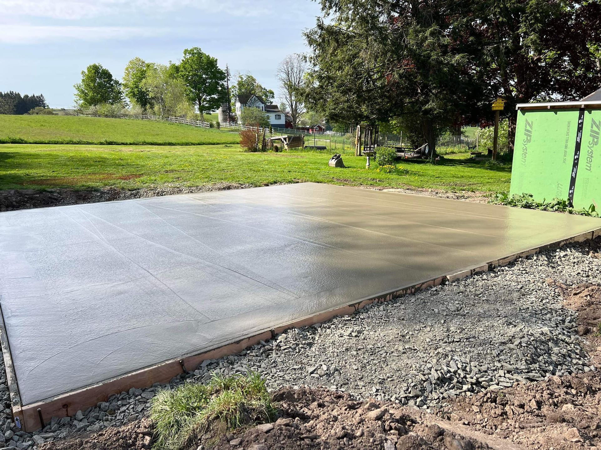 Newly poured concrete slab in a yard, bordered by gravel and wooden forms, with green siding in the background.
