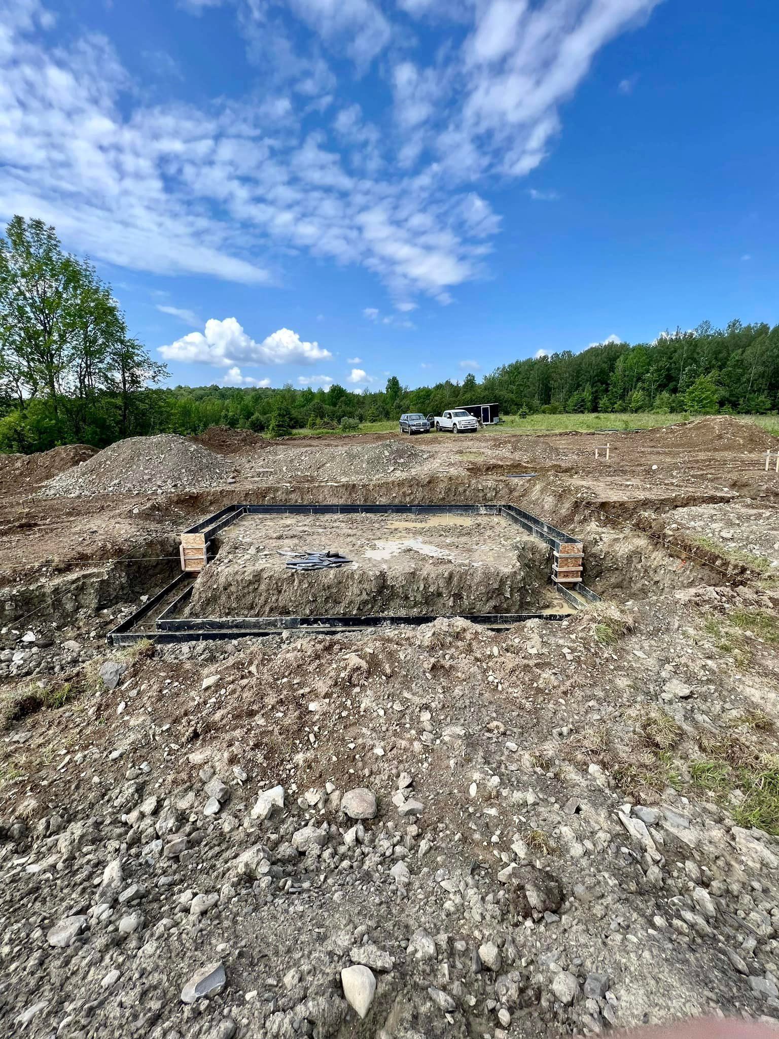 Construction site: Rectangular foundation outline dug in earth, dark pipes, bright blue sky, trees in background.