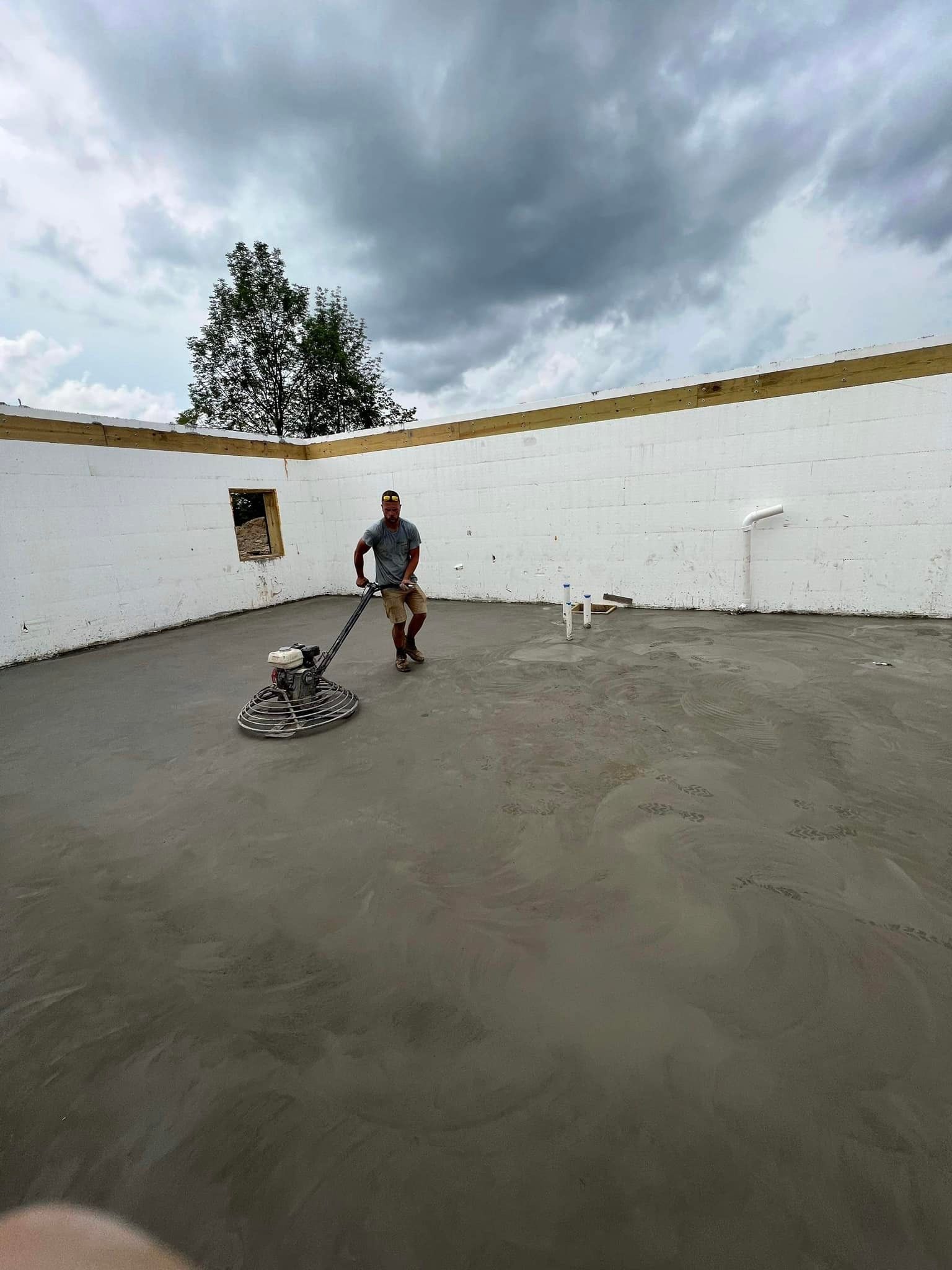 Man operating concrete grinder on a poured concrete floor within a building under construction. Cloudy sky.