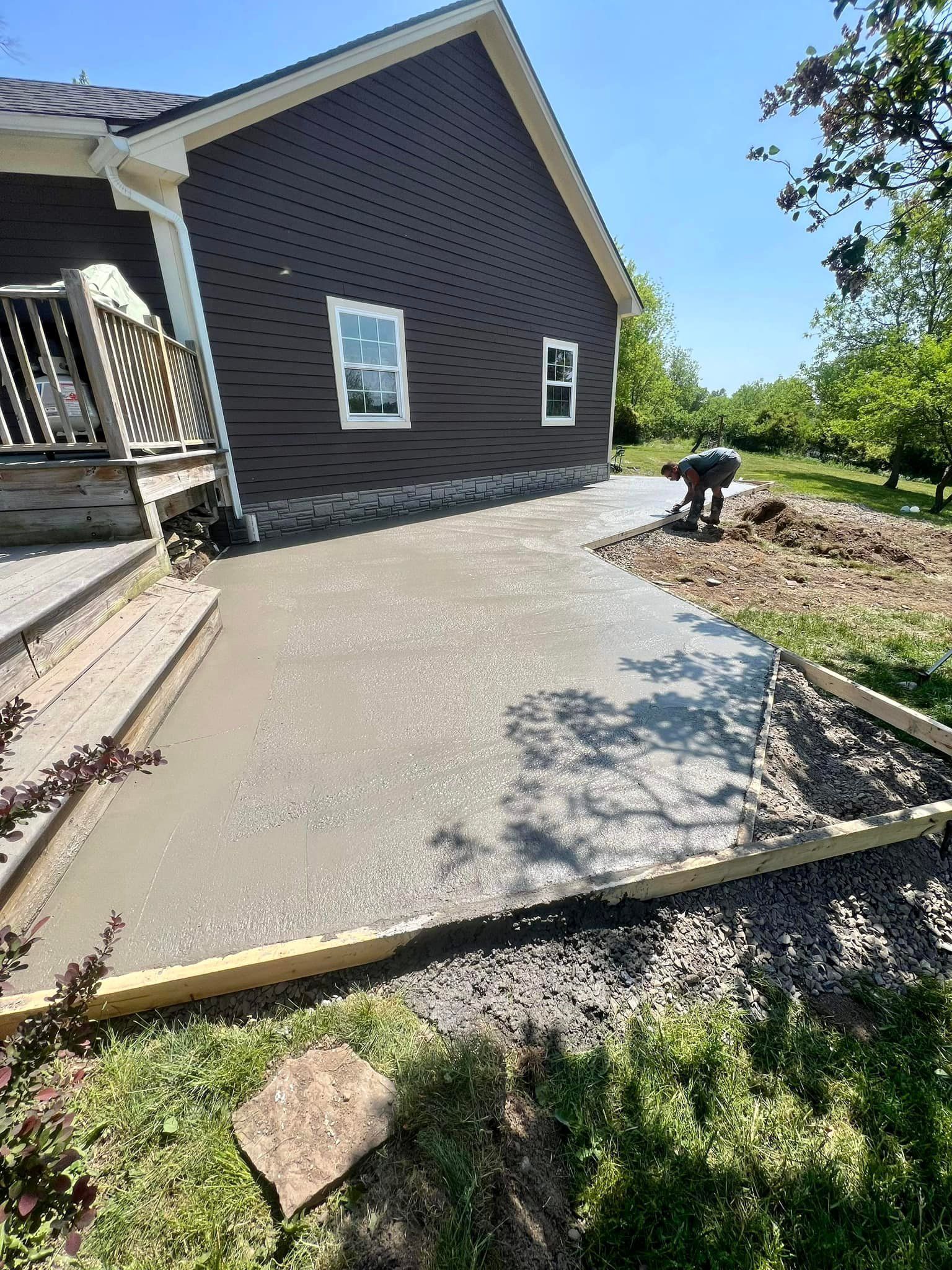 Freshly poured concrete patio next to a dark brown house with two windows and a person in the background.