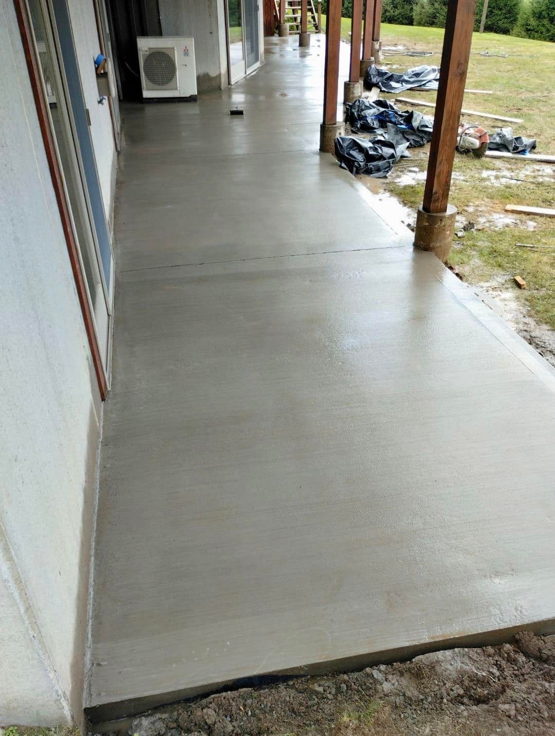 Newly poured concrete patio next to a house with an AC unit and wooden posts in the background.
