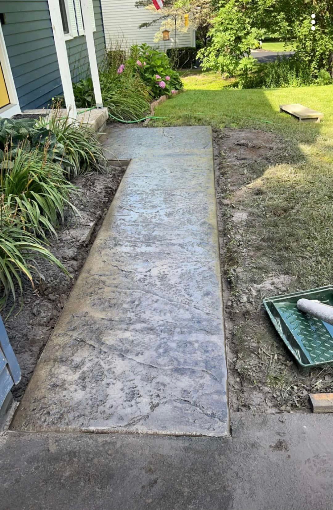Concrete walkway leading to a house, edged by dirt and grass. A paint tray and roller are visible.