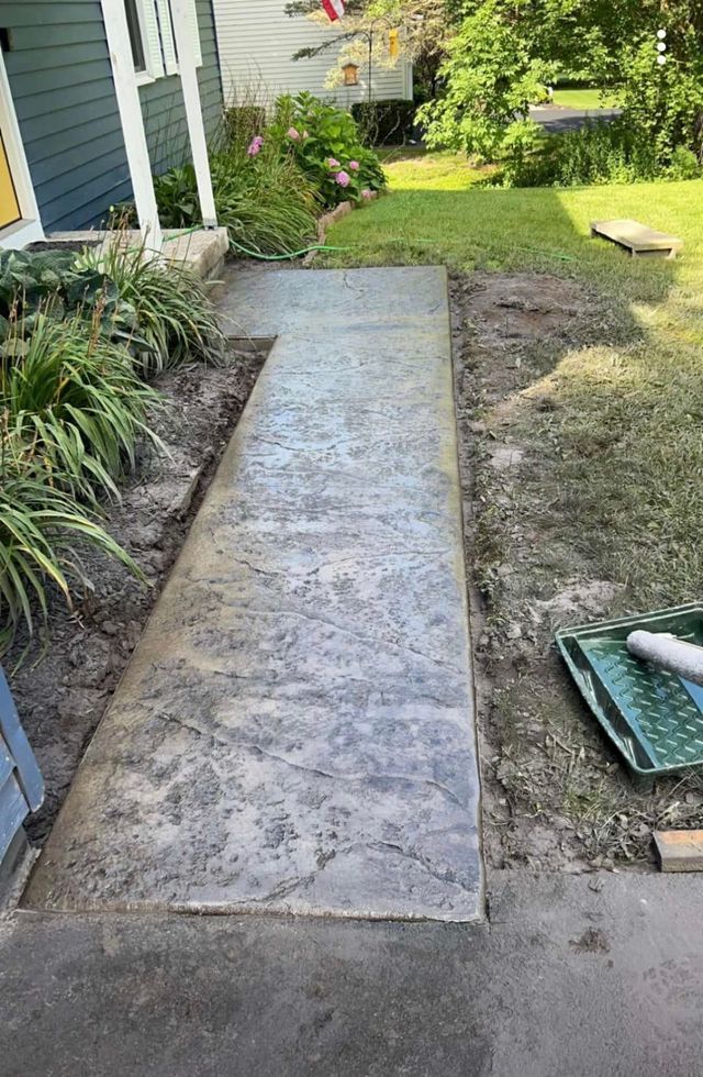Concrete sidewalk leading to a blue house, surrounded by dirt and grass.
