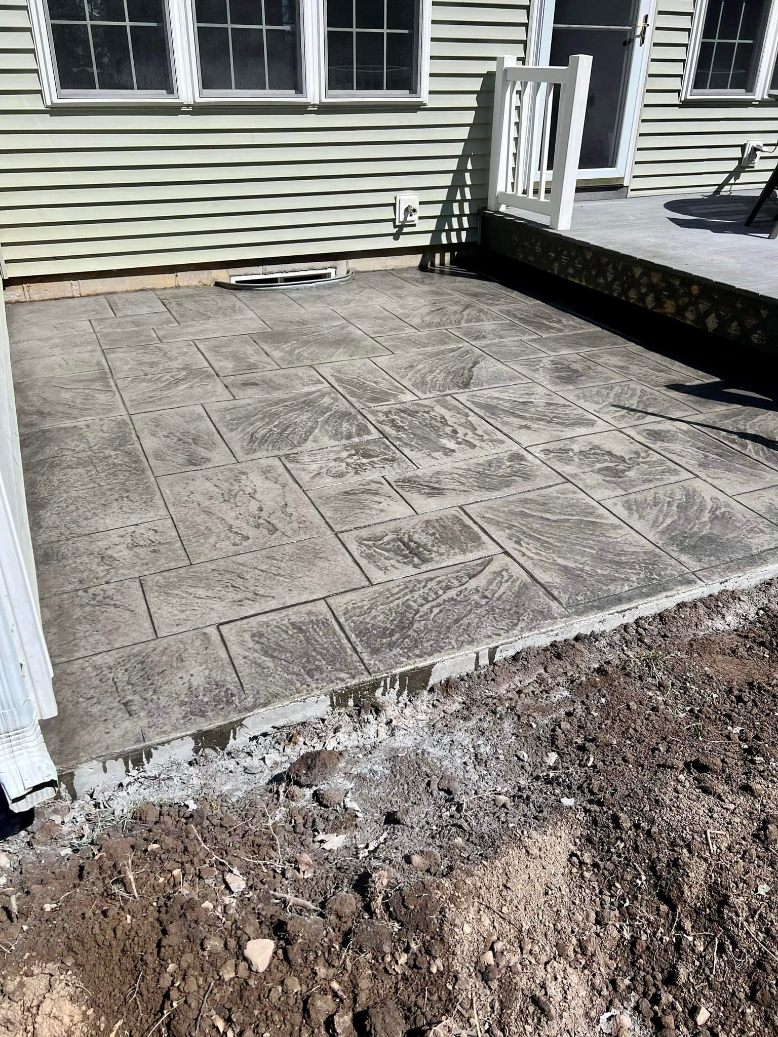 Concrete patio with rectangular pattern, next to a house with beige siding and a raised wooden deck.