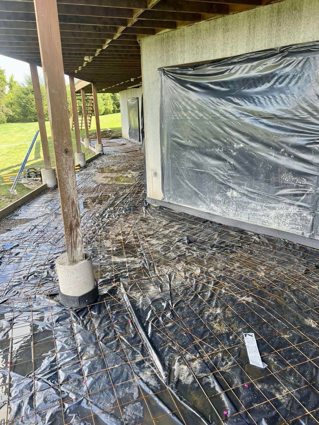 Construction site: deck with black tarp, rebar, mud, wooden supports, and building with plastic covering.