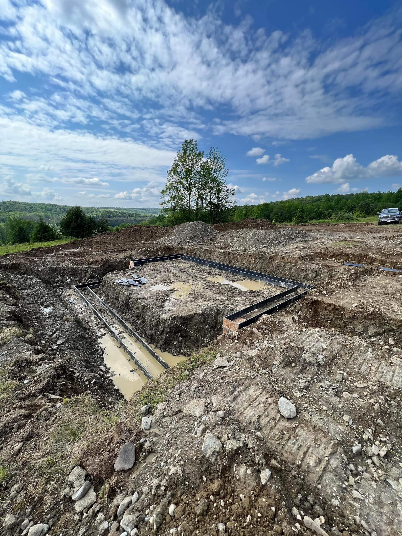 Construction site with excavated foundation. Dirt and water fill the dig. A tree stands in the background under a blue sky.