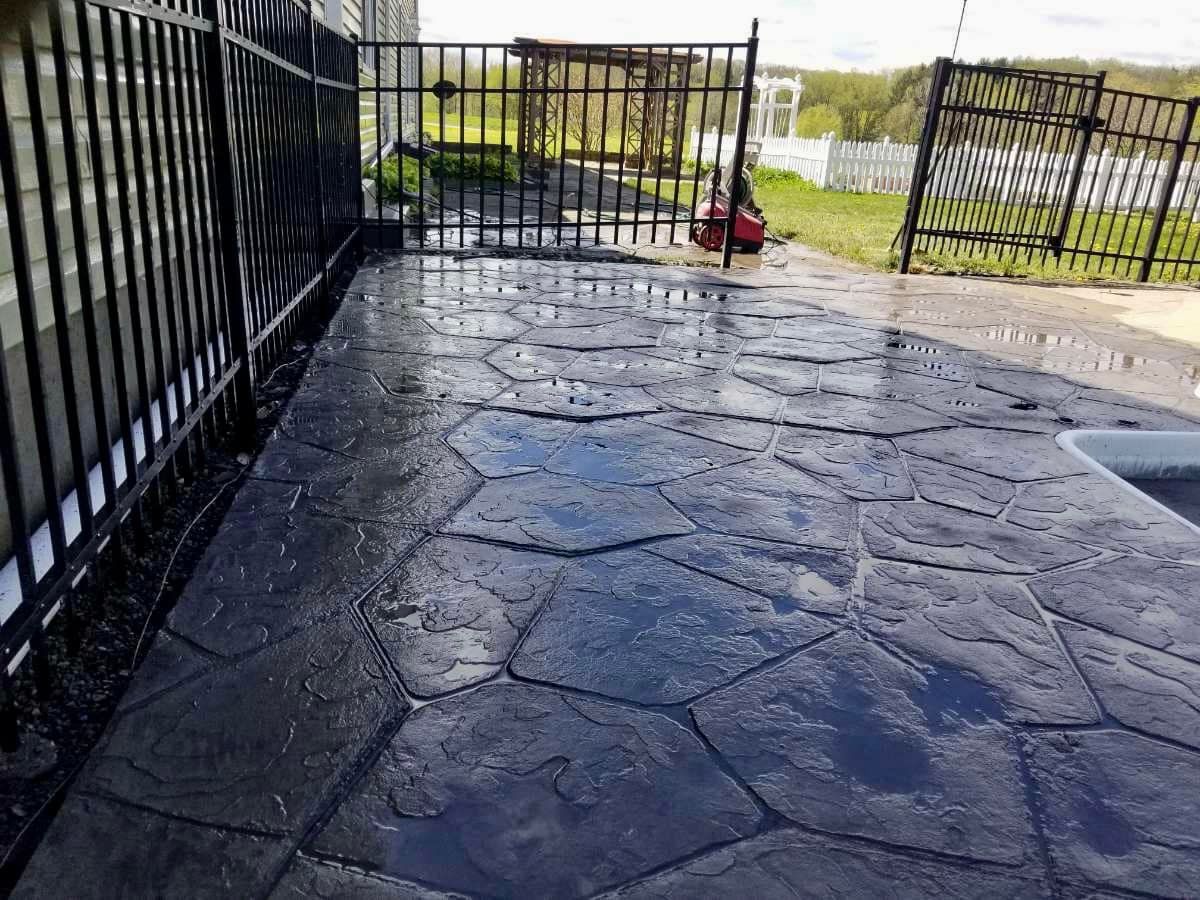 Black stamped concrete patio with wet surface, next to a black fence.