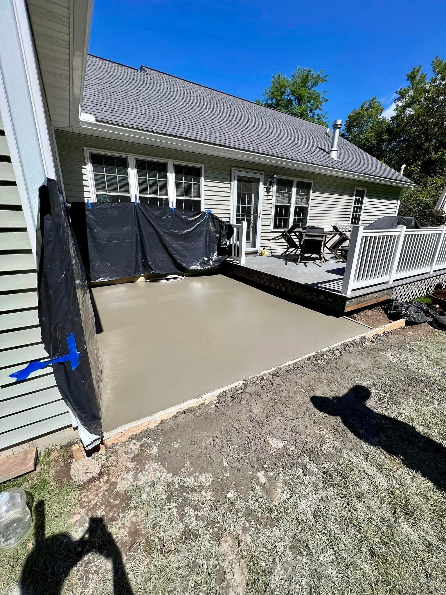 Newly poured concrete patio next to a house with gray siding and deck. Blue sky.
