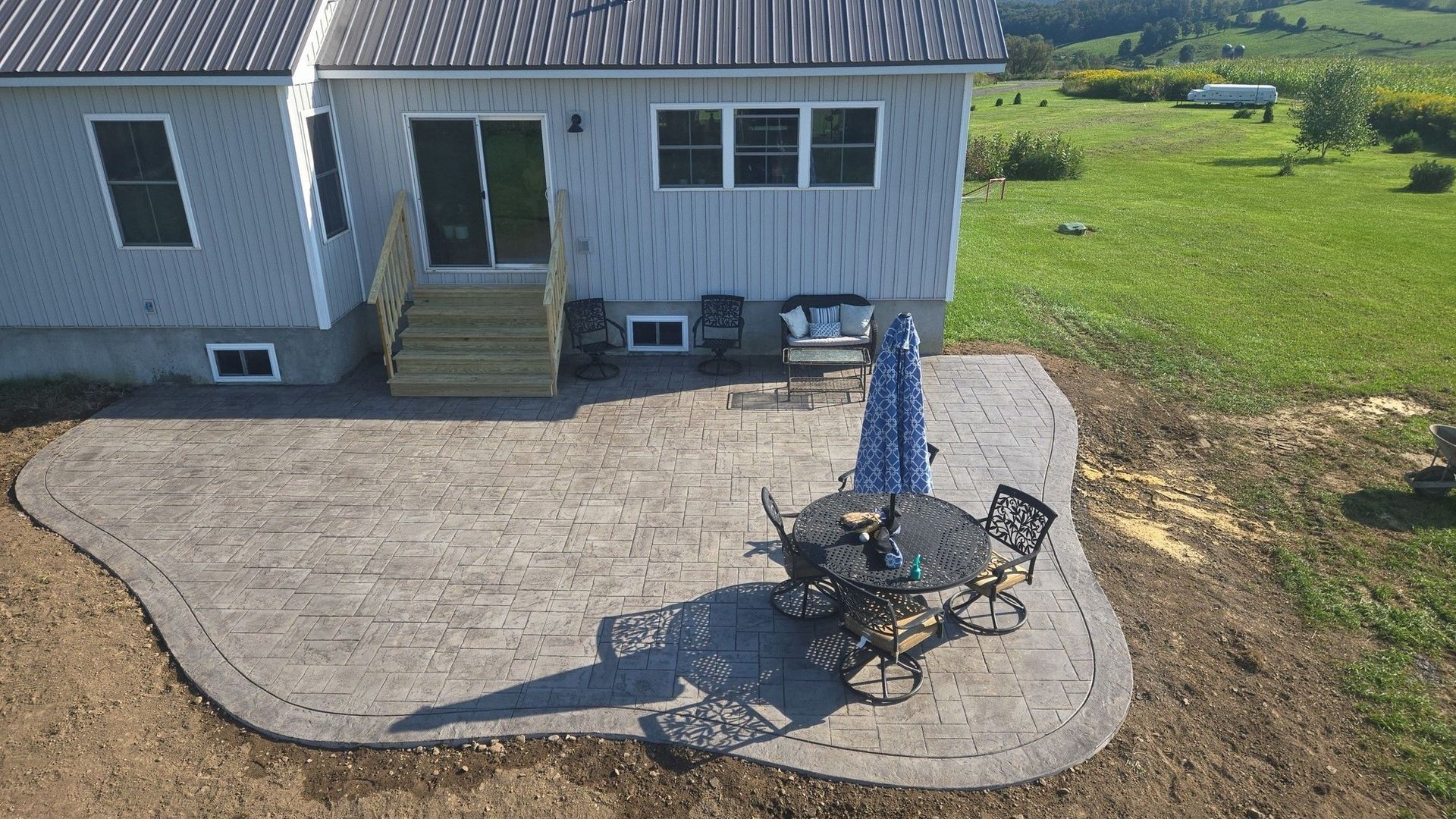 Back patio with table, umbrella, and chairs near a house with a green yard.