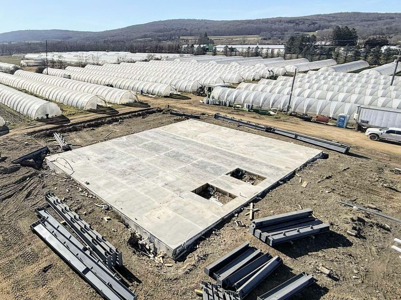 Construction site: concrete foundation with cutouts, surrounded by steel beams and greenhouses.
