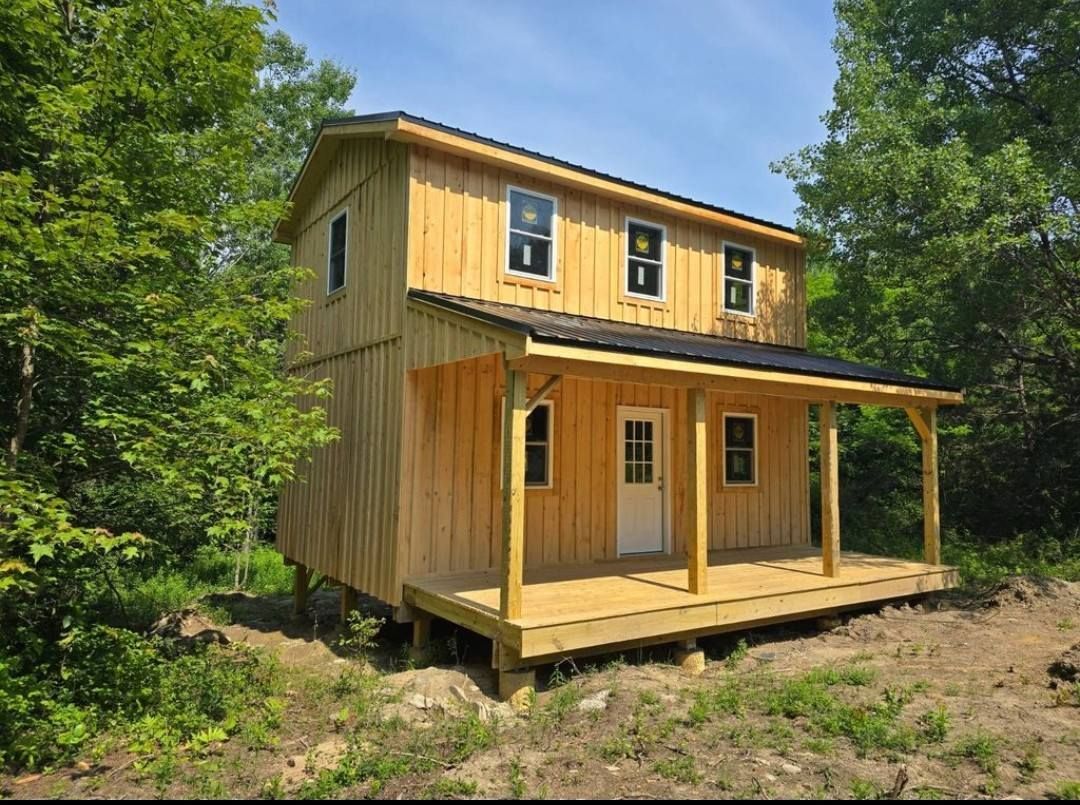 Two-story wooden house with porch, light yellow siding, black roof, and windows, set among green trees.