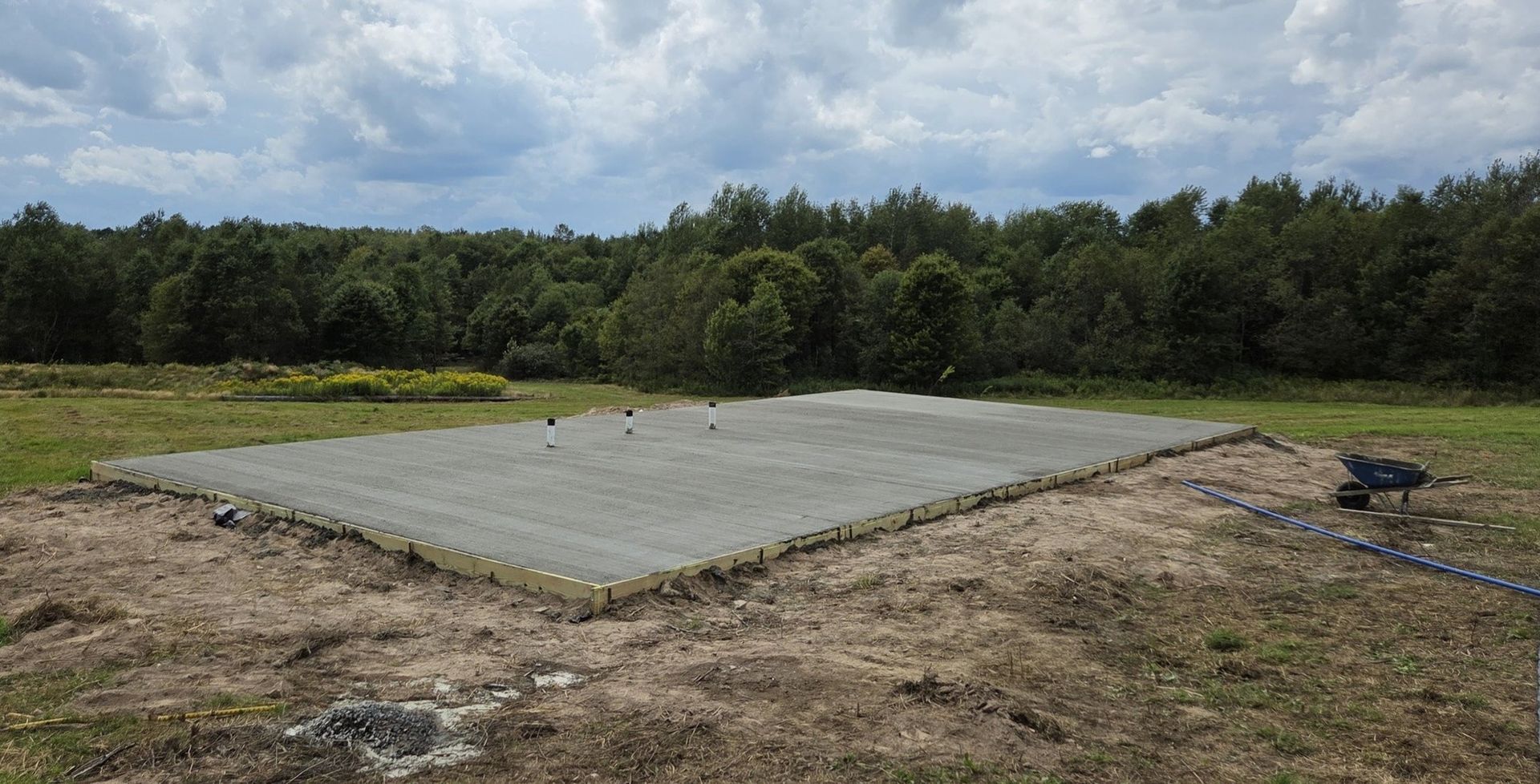 A rectangular concrete foundation with exposed rebar in a field, trees in the background, blue sky.