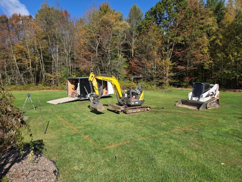 Construction site: Excavator and skid steer on grass, trailer in background, marked foundation, trees.