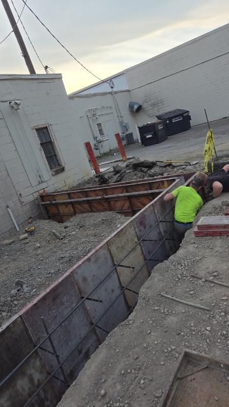 Construction worker in neon vest inspecting a deep trench beside a building and gravel lot