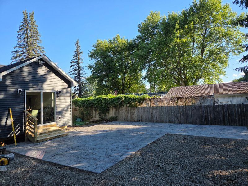 Backyard with a concrete patio beside a small gray shed and wooden fence under trees.