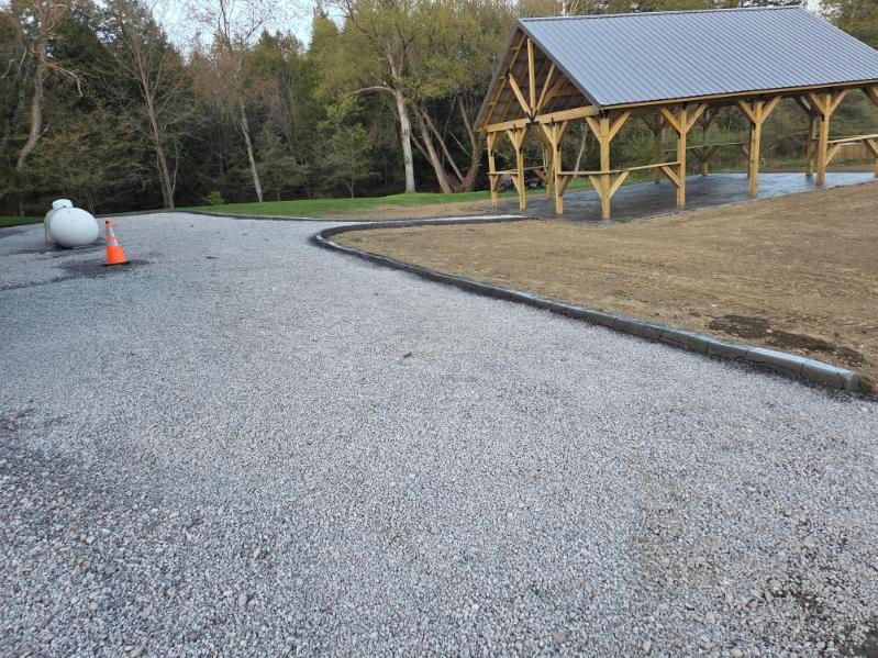 Gravel driveway beside a wooden pavilion with a metal roof, trees in the background, and a traffic cone on the left.