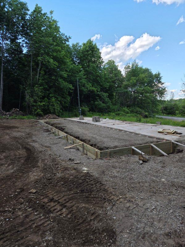 Gravel construction site with a concrete foundation slab beside a wooded area under a blue sky
