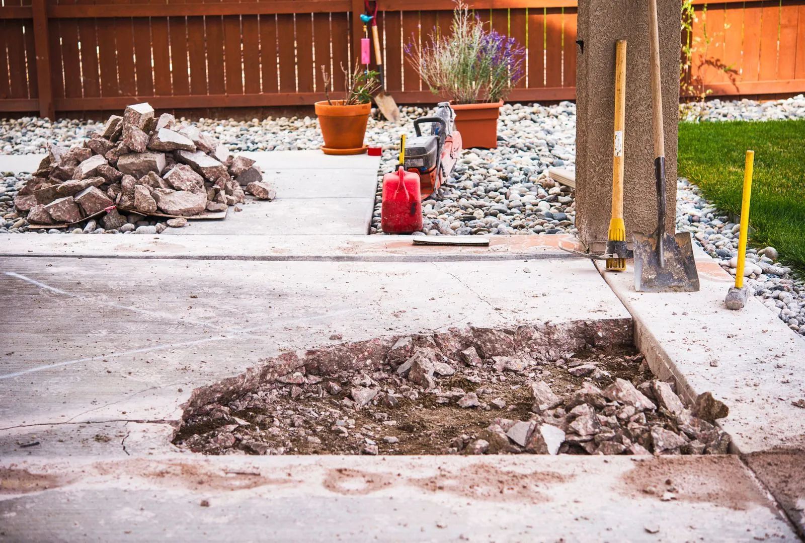 Construction site with trenches, gravel piles, potted plants, and a red fire extinguisher near a wooden fence