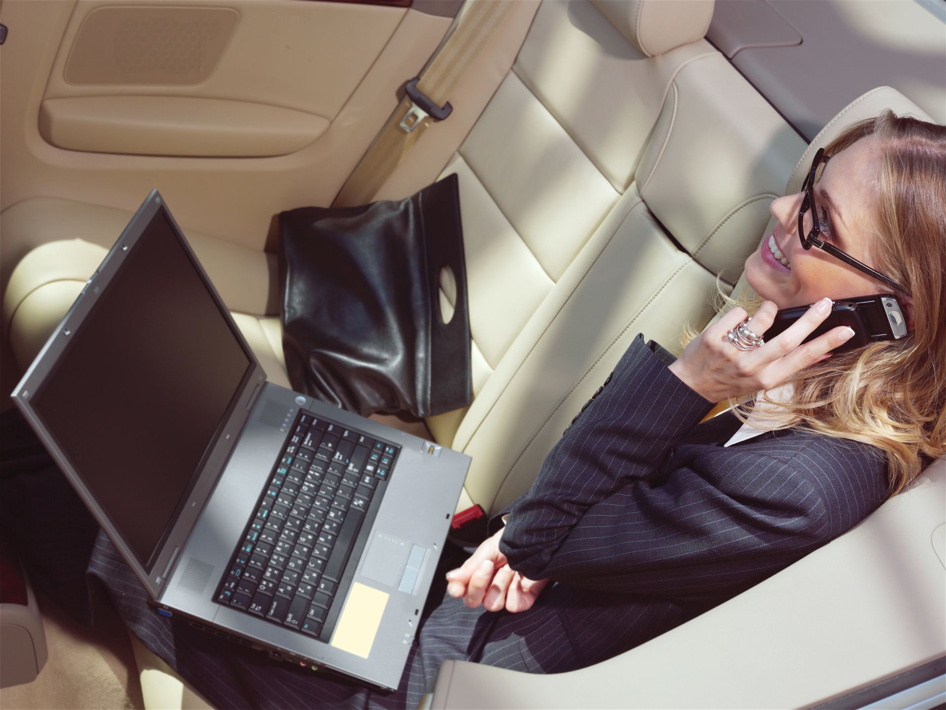 Person using a laptop and phone in a car’s back seat, wearing glasses and talking on the phone