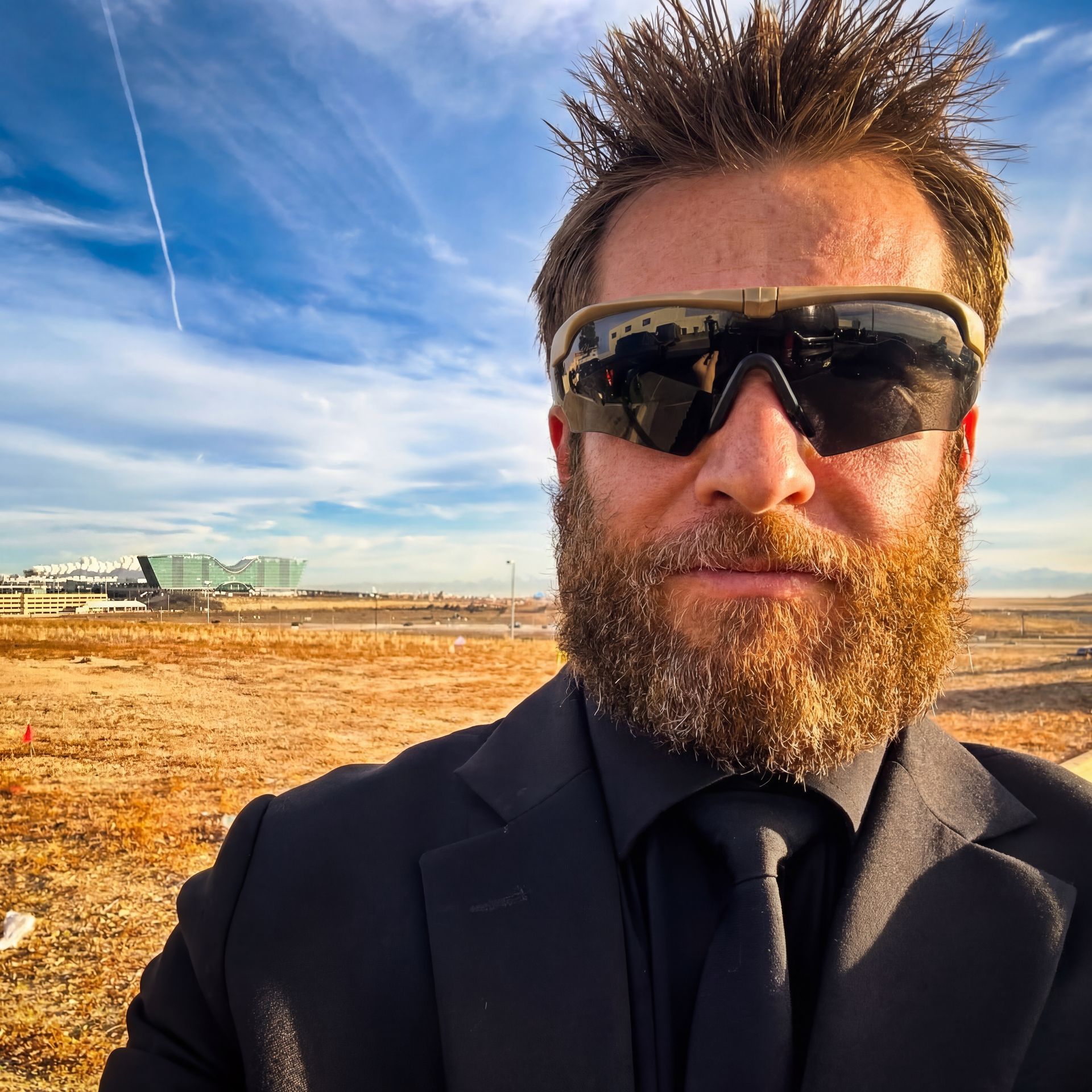 Man with beard and sunglasses in black suit, outdoors near an airport, with blue sky.