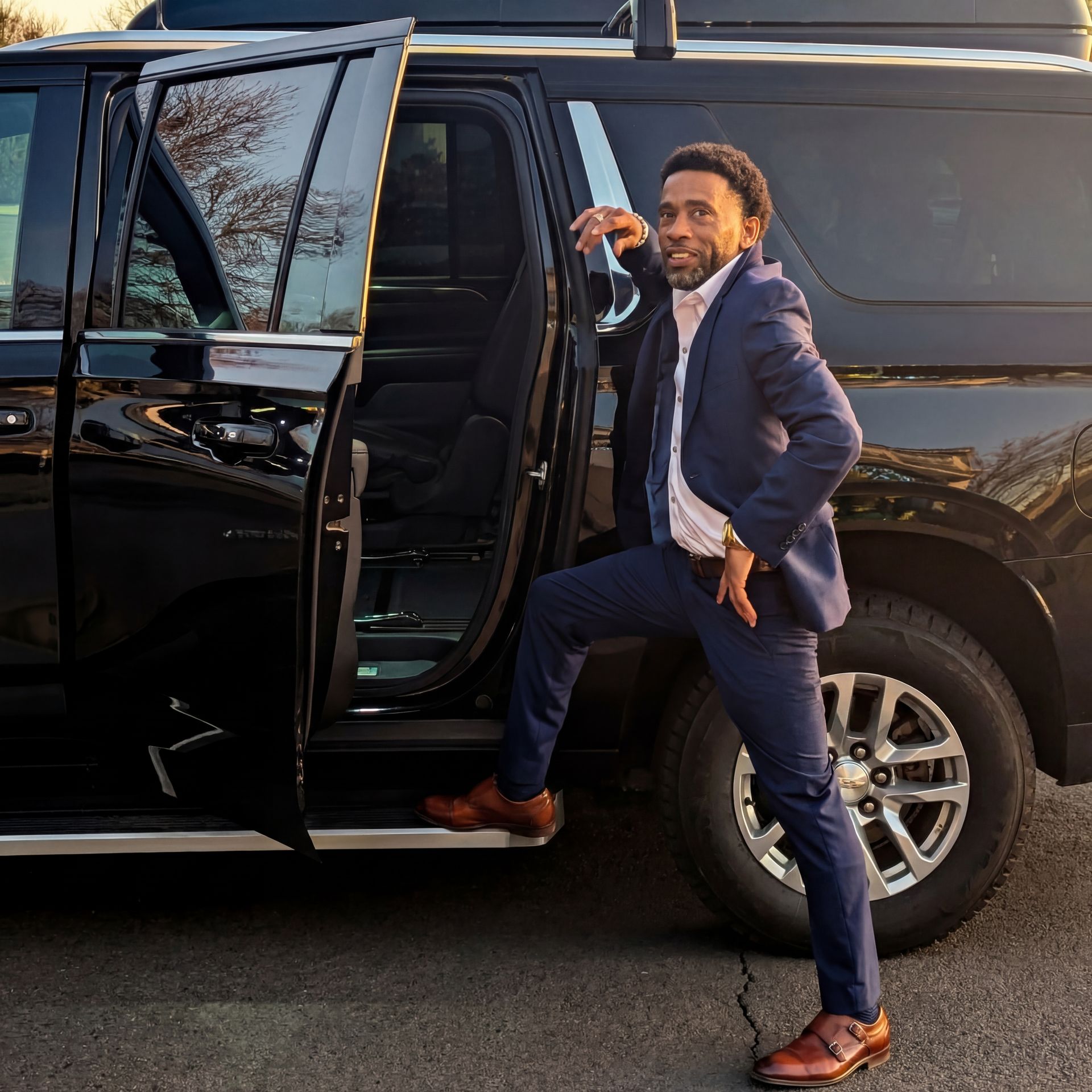 A person in a blue suit steps out of a black SUV, leaning against the open door with one hand resting on the vehicle.