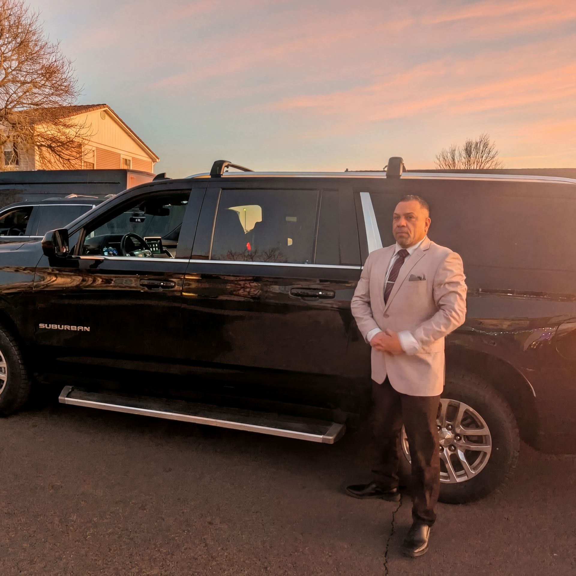 A person in a light suit stands next to a black SUV parked outdoors during sunset.
