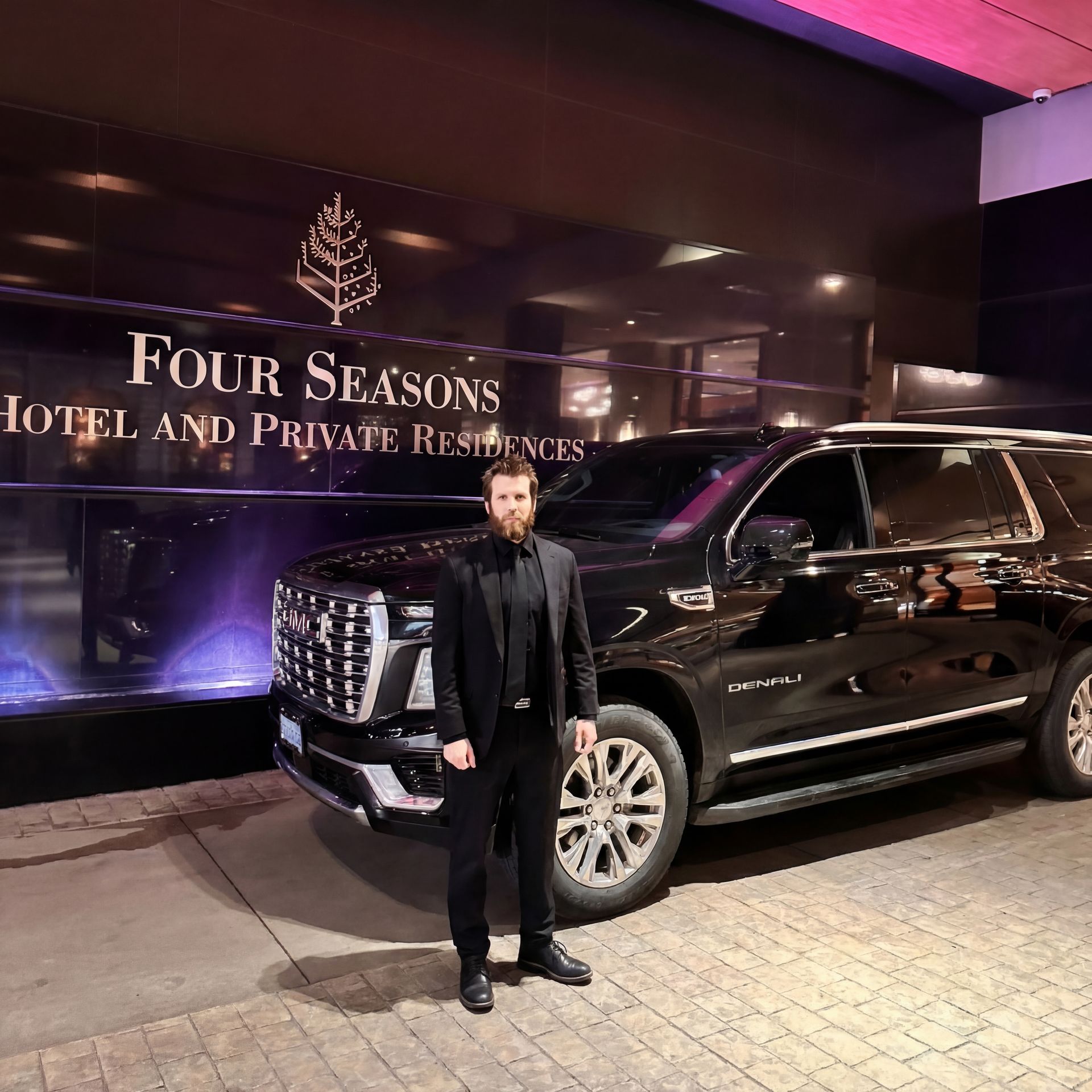 A person in a black suit stands in front of a black SUV parked at a Four Seasons Hotel and Private Residences.