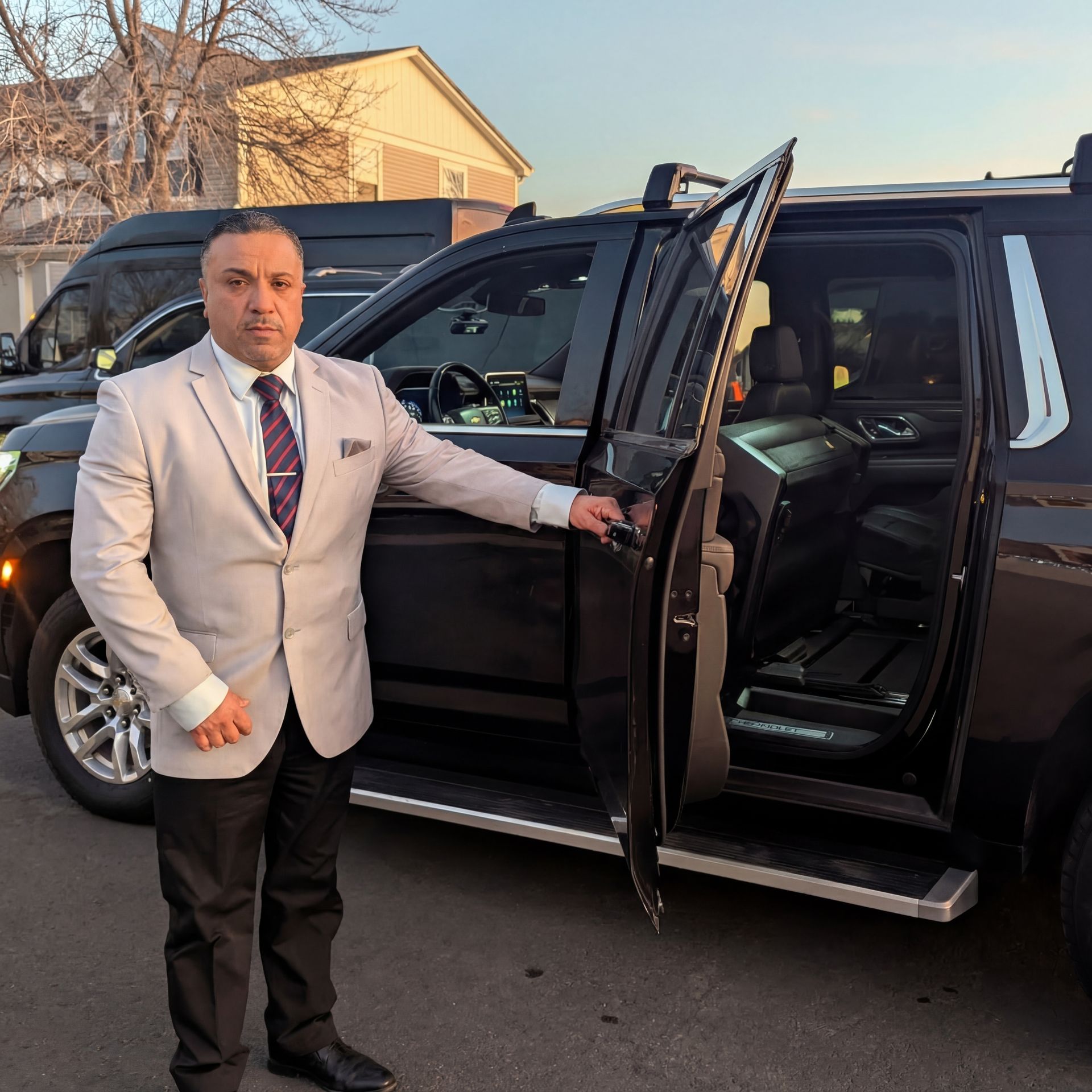 A person in a light suit and tie stands beside a black SUV with the passenger door open, parked in a residential area.