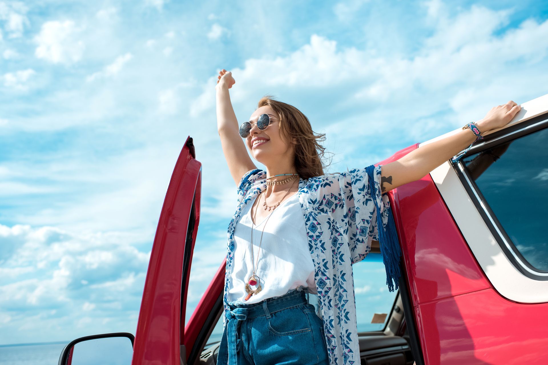 Woman standing by a red car with arms raised under a blue sky