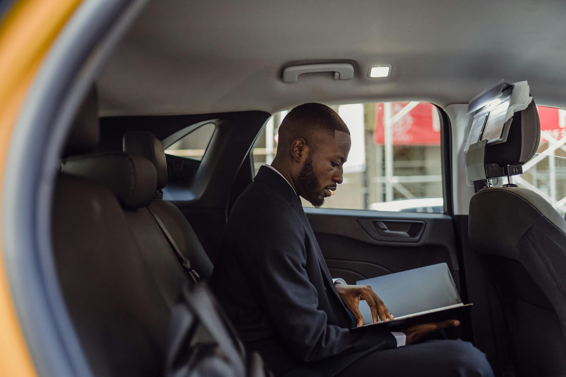 A man in a suit is sitting in the back seat of a car using a laptop.