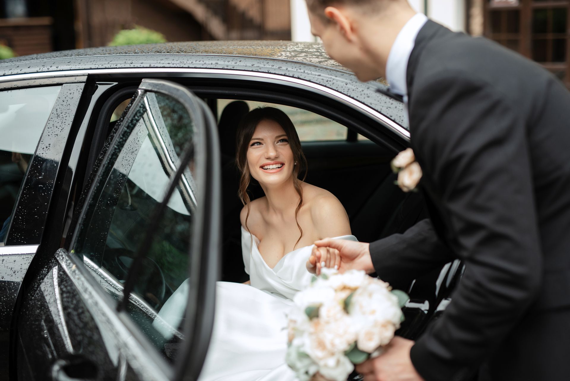Bride smiling from a car as a man in a suit opens the door, holding a bouquet