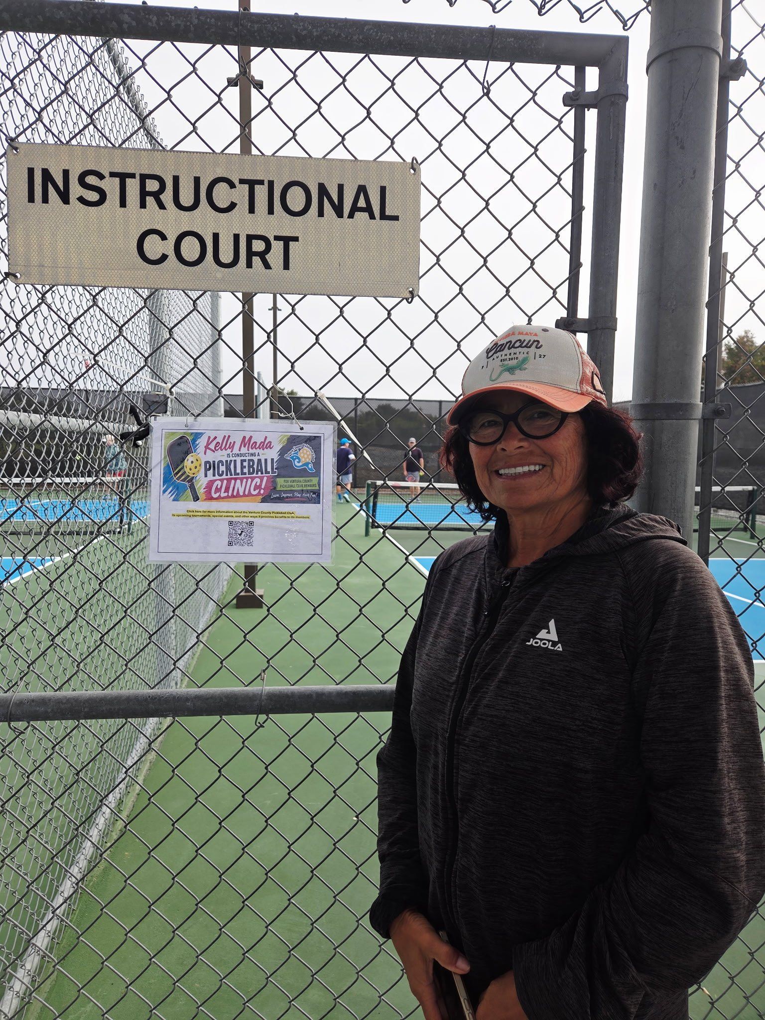 Woman smiling beside “INSTRUCTIONAL COURT” sign at a fenced tennis court.