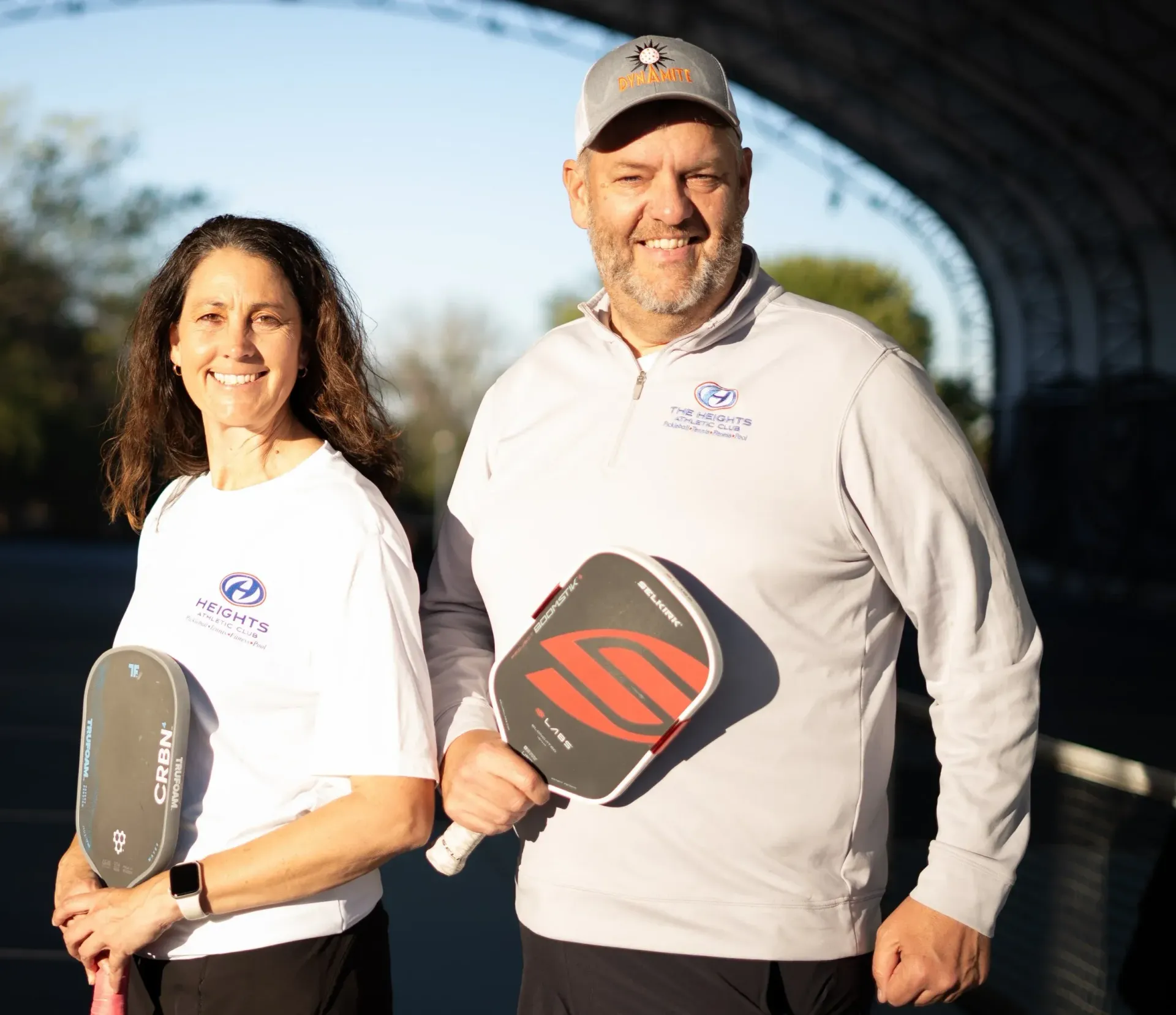 Two people holding pickleball paddles outdoors, smiling in white shirts with a blurred arched structure behind them